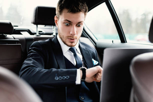 Worried stylish ceo in suit sitting on backseat of luxury car, looking on his watch, using laptop