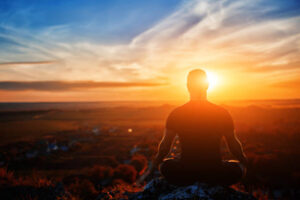 Rear view of the man meditating yoga in lotus pose on the rock at sunset. Horizontal photo. Beautiful landscape with sky and clouds like a background. Concept of the healthy and active lifestyle.