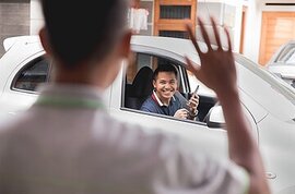 Picture of a guy from behind with his hand up waving at a smiling driver holding a phone