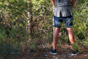 a young caucasian man wearing shorts and T-shirt seen from behind peeing in the woods