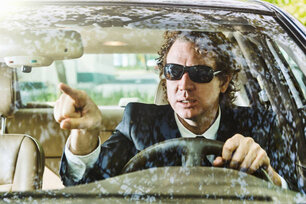 Man wearing a suit and sunglasses gestures in a direction as he drives his car.