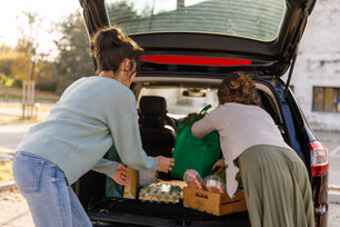 Rear view of two women loading reusable bag and a wooden crate with fresh produce, in a car trunk, after vising farmer's market.