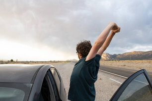 This is a photograph is of a Caucasian man in his 30s taking a break from driving through the desert in Nevada to stretch his arms overhead while standing outside the car.
