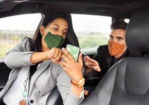 Female taxi driver showing to the passenger the map on the smartphone, both wearing protective face mask during pandemic.