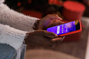 Close up shot of unrecognizable young Black woman sitting by the Christmas tree and rating her experience after doing online shopping via smart phone.