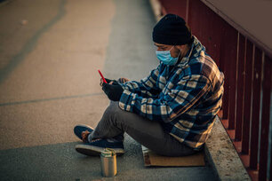 Homeless man with protective mask sitting on the bridge with donation can and using mobile phone
